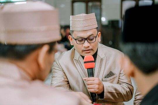 Close-up of an Asian Muslim man wearing a traditional peci and suit, holding a microphone during an Islamic wedding ceremony. Ijab qabul concept, solemn religious event in Indonesia.
