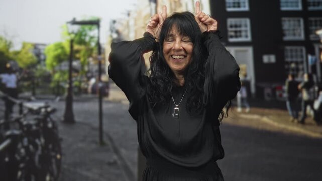 Woman senior hispanic smiling, wearing pendant necklace and black dress, hands making horns gesture on a cobblestone street with bicycles and buildings in view; playful joy.