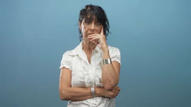 Woman senior hispanic with hand to mouth and crossed arms wearing white blouse and silver cuff in studio; contemplation resilience.