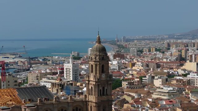 Aerial shot rising forward over Malaga cathedral with tilt down revealing dense city rooftops and coastal harbor in summer midday