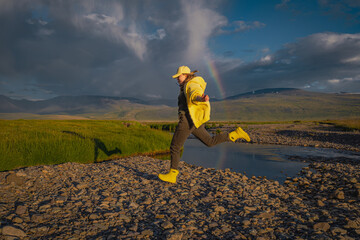 Fototapeta premium Energetic young woman in yellow boots jumping over a stream under a rainbow and dramatic storm clouds during golden hour sunset, representing joy, hope, and freedom in a wild mountain landscape