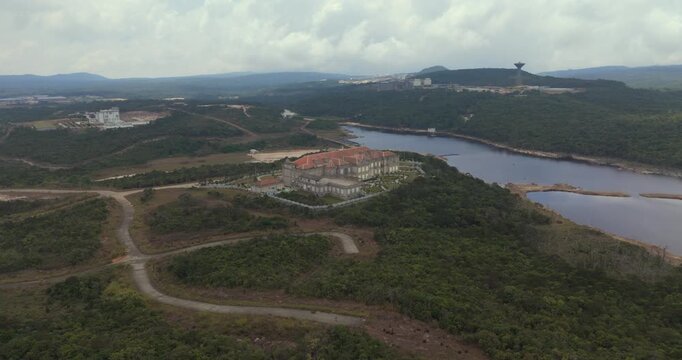 Forward  aerial view of a grand red roofed mansion on Bokor Mountain, Cambodia, overlooking the dark waters of Bokor Lake and featuring a distant water tower on a forested mountain ridge.