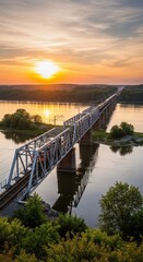 Obraz premium Stunning sunset over a long railway bridge spanning a wide river, with lush green trees on the banks and a vibrant orange sky reflecting on the water.