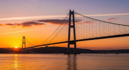 Obraz premium Stunning sunset over a majestic suspension bridge spanning a wide body of water, with vibrant orange and yellow hues reflecting on the calm surface.
