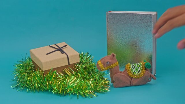 Hand placing camel toy into holiday scene with gift box and silver book against teal backdrop