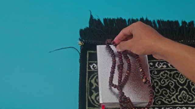 Hand places wooden prayer beads onto Quran resting on black prayer rug over teal background