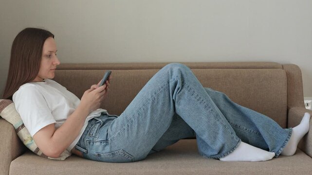 Side view footage. Woman with long brown hair relaxes on a couch while using a smartphone, wearing a white t-shirt and blue jeans in a minimalist indoor setting