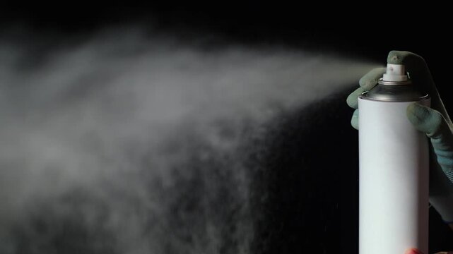 Hand wearing work glove spraying white paint from aerosol canister against black background.