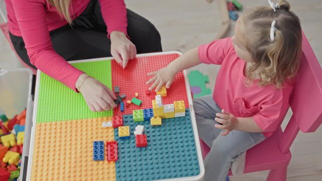 Woman guiding toddler placing colorful interlocking blocks with hands on activity table in playroom with foam mats and toy bin visible; bonding play.
