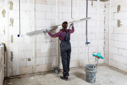 A woman in overalls checks the plastering guides on a wall of white blocks with a rule. A professional female construction worker at work on an interior renovation site