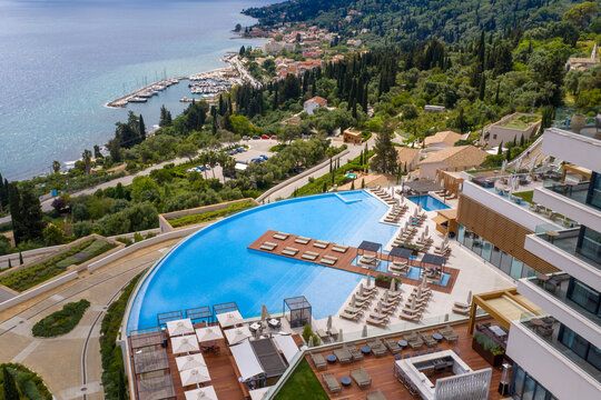 Aerial perspective of a modern luxury hotel in Corfu featuring a large infinity swimming pool and sun loungers. Scenic view of the Greek coast, Benitses harbor, and the Ionian Sea