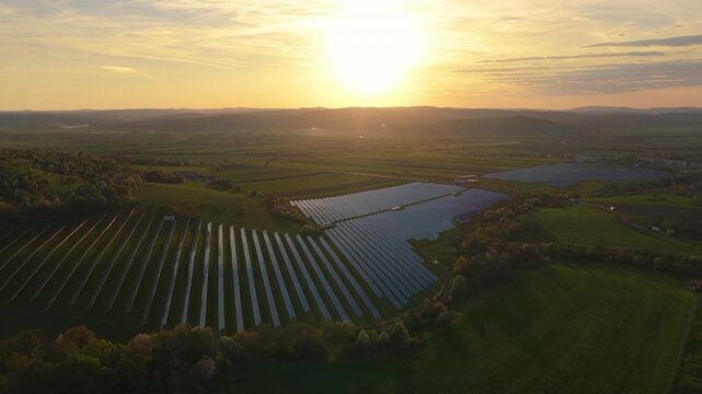 Stunning aerial view of a massive photovoltaic solar park on rolling hills surrounded by blossoming spring trees at sunset, high altitude lateral tracking drone shot, graded