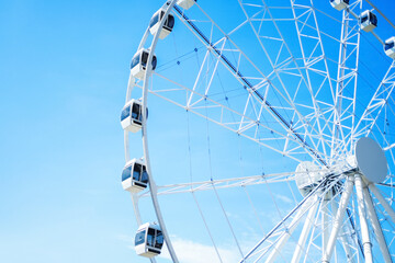 A large white Ferris wheel with modern enclosed gondolas set against a vibrant, clear blue sky. A minimalist and bright scene capturing summer fun and amusement park vibes. © Kyrylo