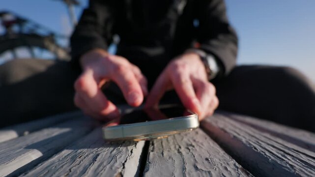 A man taps and swipes on a smartphone lying flat on a weathered wooden bench. Close up shot of hands interacting with the touchscreen device outdoors under bright sunlight.