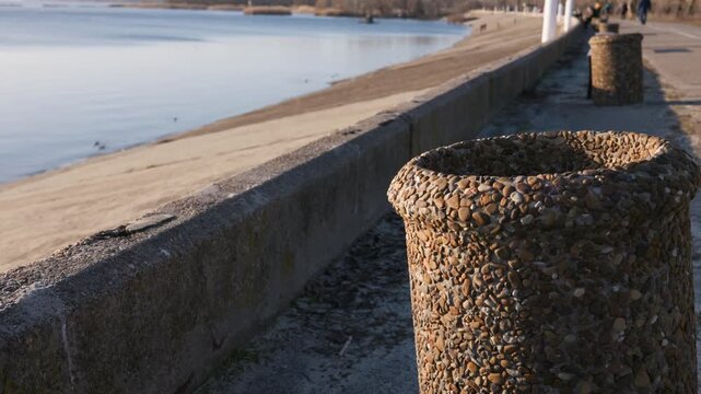 A stone trash can stands on a paved waterfront promenade beside a river. A concrete embankment separates the walkway from the calm water. Blurred pedestrians stroll in the distance.