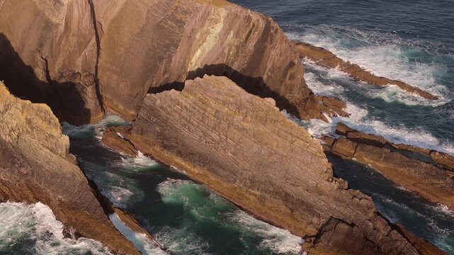 Witness the immense power of nature as dynamic ocean waves continuously crash against the strikingly beautiful, stratified rock formations of a rugged coastline at sunset.