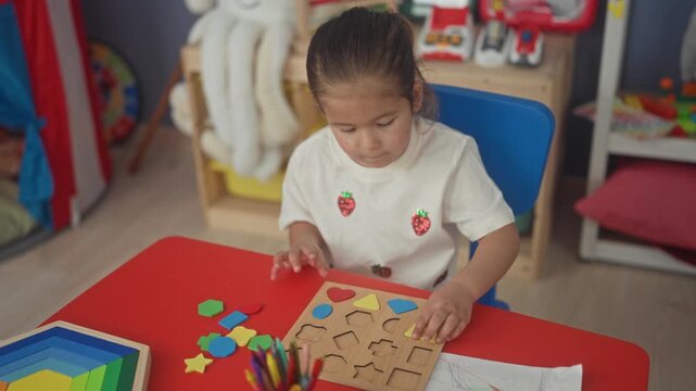 Little girl placing a red wooden shape into a shape sorter board with her hand at a building playroom table with rainbow stacker and colored pencils visible; concentration learning.