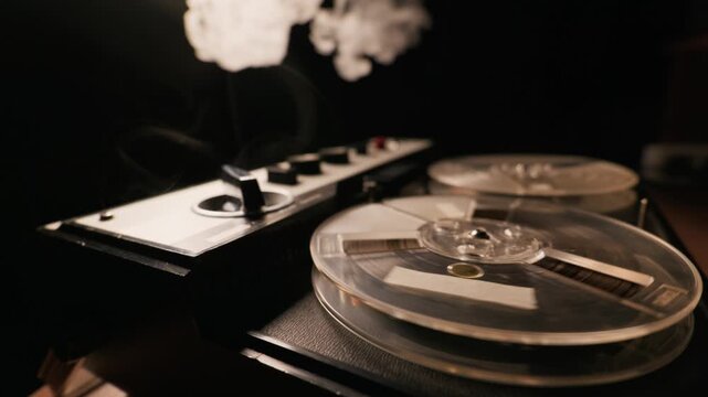A vintage reel-to-reel tape recorder spins continuously in complete darkness. Thick white smoke billows over the mechanical spools and magnetic tape under cinematic lighting.