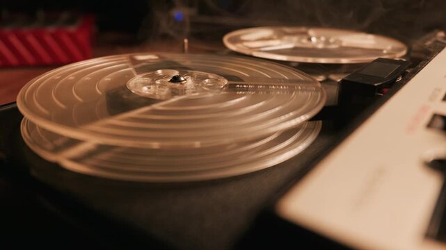 A vintage analog reel to reel audio tape recorder plays magnetic tape. Clear plastic spools spin continuously on the dark equipment console. Light smoke drifts over the machine.