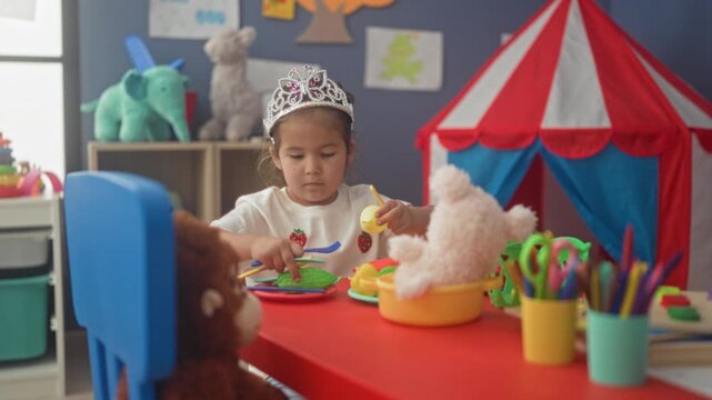 Girl wearing tiara feeds stuffed bear with toy fork at red play table in a building playroom; concentration imagination.