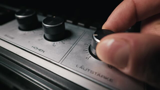 Human fingers carefully turn the master volume control knob on a vintage stereo amplifier. The brushed silver metal panel features German text labels indicating bass and treble adjustments.