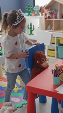 Hispanic girl wearing tiara gently places stuffed monkey on blue chair in bright playroom inside building; playful imagination.