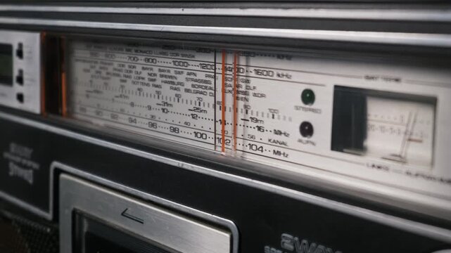 Close up view of a vintage analog radio receiver dial interface. The glass panel displays multiple broadcast frequency bands and European city names. Red tuning needles rest on the scale.