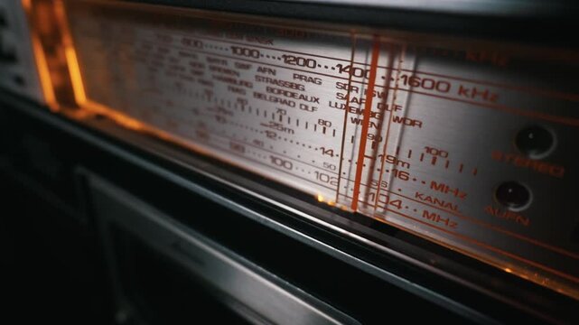 Close-up of an antique radio tuner dial glowing with warm orange light. The metallic panel displays various frequencies in kilohertz and megahertz. A red tuning needle rests on a station.