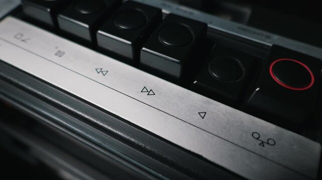 A finger presses a mechanical button on a vintage tape deck in macro view. The silver metallic panel features engraved media symbols in sharp detail. Soft light highlights the analog action.