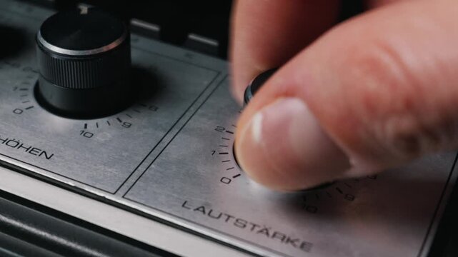 Fingers gently turn the black volume knob on a vintage silver audio amplifier with shallow depth of field. Concept of adjusting music sound levels and using classic analog retro equipment.