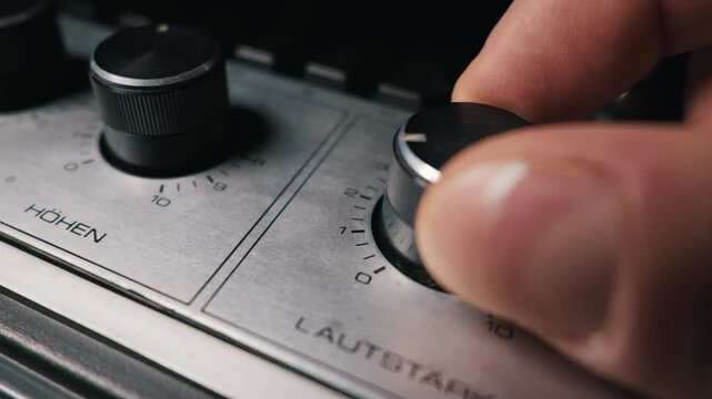 A person fingers carefully turn the metal volume dial on a vintage silver audio amplifier with German text. Audio control concept representing sound adjustment on classic electronic hardware.