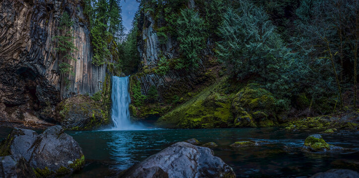 Panoramic ground view of Toketee Falls cascading into a turquoise pool surrounded by basalt columns and a lush evergreen forest in Oregon, USA.
