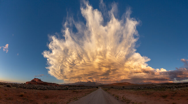 View of a dirt road leading towards a massive dramatic cloud formation over the desert landscape at sunset, Utah, United States.