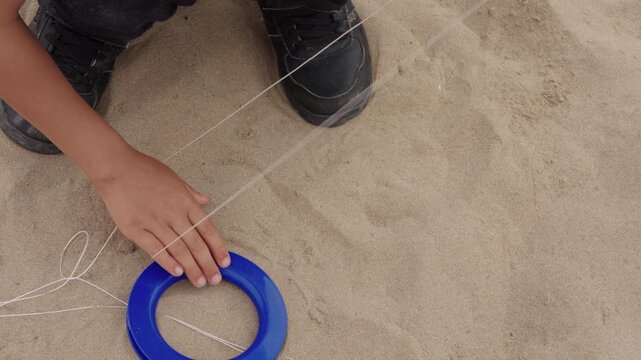 Close up view of child hands winding kite string on a spool while sitting on sandy beach surface.