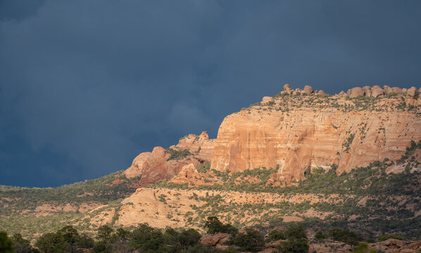 View of sunlit red rock cliffs and desert vegetation under dramatic dark storm clouds in Moab, Utah, United States.