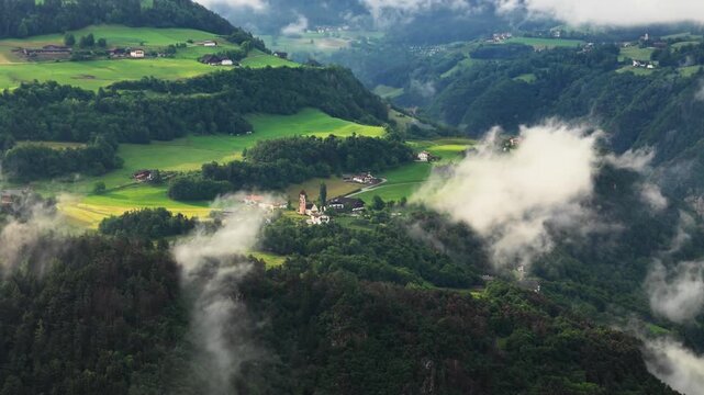 Aerial view of small alpine village surrounded by green hills and drifting fog in South Tyrol, Dolomites, Italy. Rural mountain living with cloudy weather