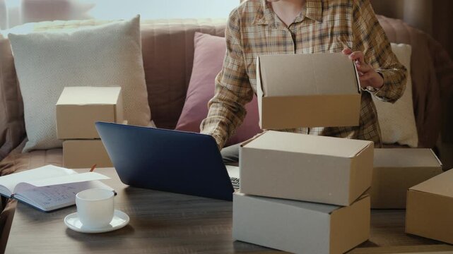 Woman entrepreneur working on laptop at home office preparing cardboard parcel boxes for shipment for online store ecommerce startup.