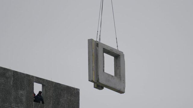 Construction scene unfolding. Steel cables hold precast panel near building facade. Worker observes as precast concrete panel is carefully hoisted into position