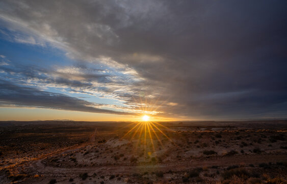 View of a dramatic sunburst over the desert landscape with scrub brush and dark clouds in Moab, Utah, United States.