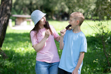 Naklejka premium Happy mother and son enjoying chocolate ice cream on a stick while walking in a sunny summer park. Young woman in a hat and a school-age boy eating popsicles, laughing and having fun together