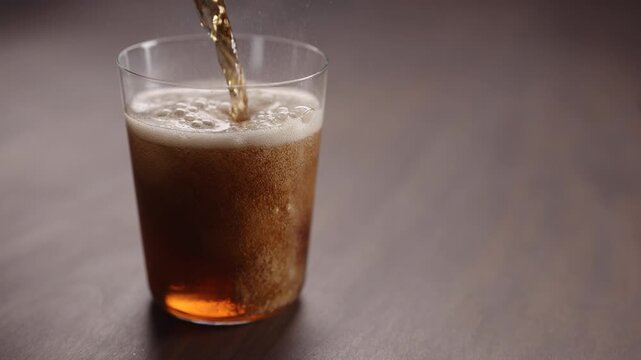 Slow motion man pour cola over ice cubes into tumbler glass on walnut table with copy space