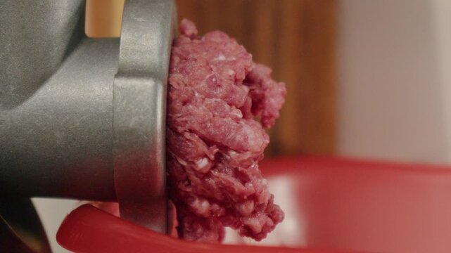 Meat grinding process. Freshly minced beef flowing into bowl in warm kitchen setting. Home cook carefully processing raw beef through grinder for burgers and sausages