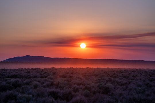 View of the sun setting over a desert landscape with sagebrush and distant mountains under a hazy orange sky in Moab, Utah, United States.