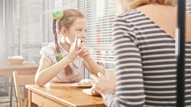 Girl with pigtail eats sweet cakes in the cafe, talks with her mum and wipes her hands with a napkin