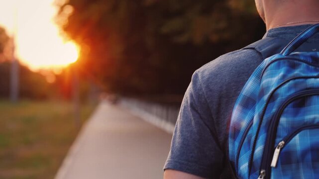 Young man walking with a blue backpack towards golden sunset in the park.