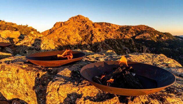 Ancient forged iron signal fire pans with burning charcoal and glowing flames resting on rocky mountain cliffs during golden hour sunset, cinematic historical landscape with rugged stone peaks.