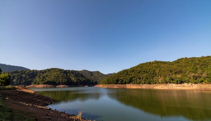 Fototapeta premium Serene lake surrounded by lush green hills under a clear blue sky.