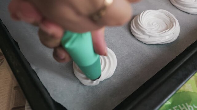 Hand of a cook squeezes white meringue mixture from a green piping bag onto a baking tray with parchment paper. Concept of homemade dessert preparation, baking process, and culinary art.