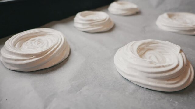 White meringue swirls sit neatly on a baking sheet lined with parchment paper. This shot captures the glossy texture of fresh homemade dessert preparation in a bright kitchen setting.