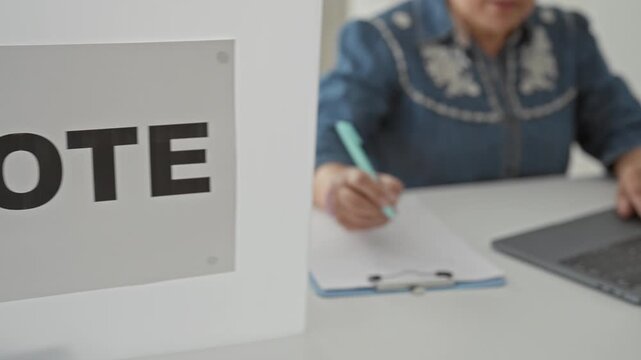 Woman with short gray hair smiles and taps laptop touchpad while holding pen at voting table in a building; civic pride.